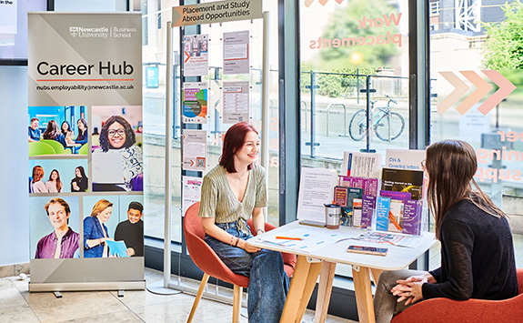 A student with light skin tone and red hair seated at a table in the Career Hub, discussing employability resources with a consultant. Brochures and a Career Hub are banner in the background.