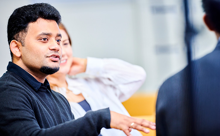 A medium-skinned male student in a black jumper gestures while speaking during a discussion, with other students listening in the background.