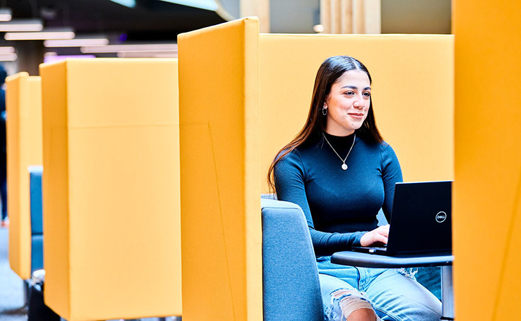 Young woman with light-medium skin tone, long brown hair, and a black top working on a laptop in a yellow study booth.