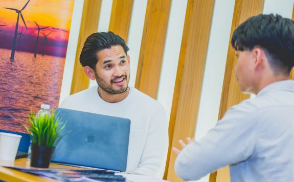 Two students converse at a networking event. One, with a laptop open, listens attentively, while the other gestures as he speaks. A poster of wind turbines at sunset and a small plant are visible.