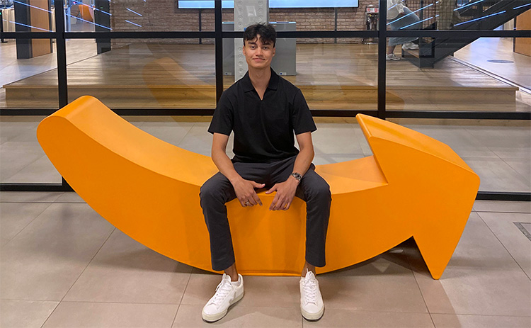 A male student with light to medium skin tone, wearing a black shirt, grey trousers, and white trainers, seated on an orange Amazon logo-shaped bench in a modern office setting.