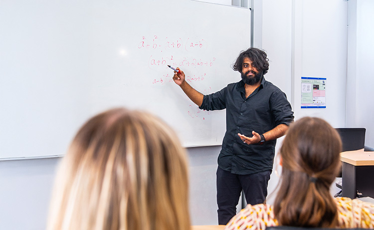 A medium-dark skinned male student wearing a black shirt is presenting mathematical equations on a whiteboard to students. The backs of two students are visible in the foreground.