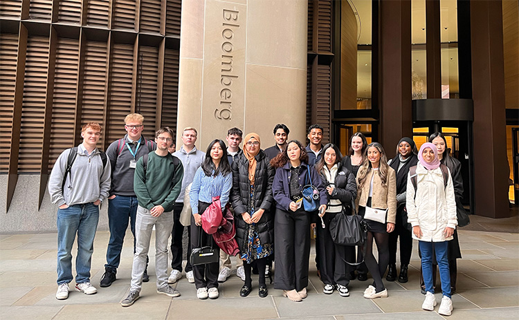 A group of students visiting Bloomberg headquarters during the student trek. They are standing outside the building, in front of a large pillar with the Bloomberg logo.