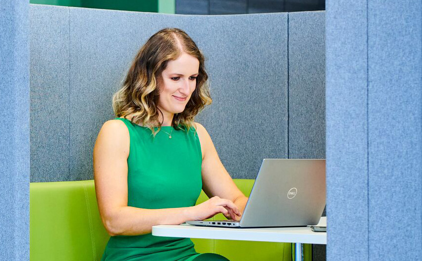 A light-skinned woman with shoulder-length wavy hair, wearing a green dress, working on a Dell laptop in a modern study booth with grey fabric walls and a green seat. The setting appears professional and comfortable.