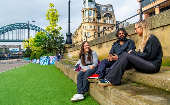 Three students, one with light skin and dark hair, one with medium-dark skin and shoulder-length curly black hair, and one with light skin and long blonde hair, sitting on steps outdoors by the Newcastle Quayside with the Tyne Bridge in the background