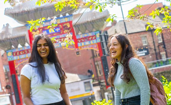 Two light-skinned women stand outdoors, laughing together in front of the Chinatown arch in Newcastle.