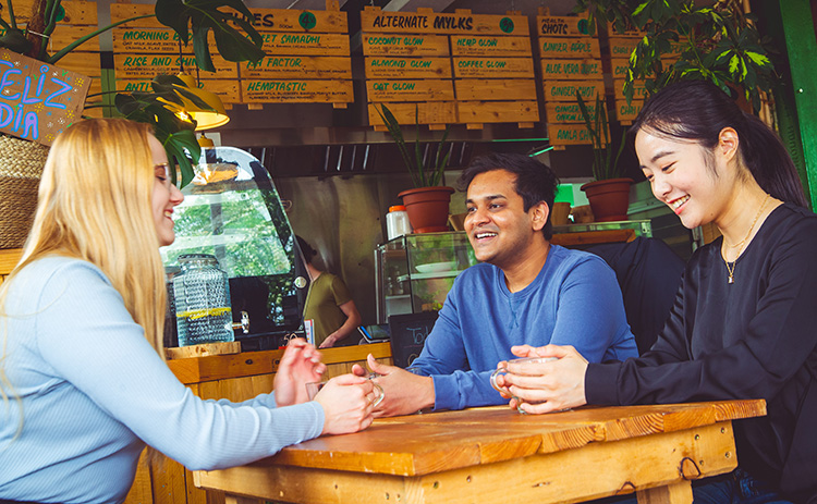 A diverse group of three students engaged in a friendly conversation at a vegan cafe, seated around a wooden table, with menu boards and potted plants in the background.