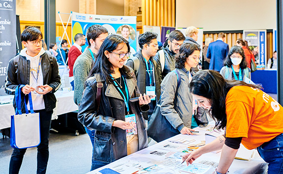 A group of students, including several light-skinned and medium-skinned individuals, engaging with a representative at a careers fair booth. One student is smiling while holding a leaflet. The event setting includes promotional banners and informational tables.