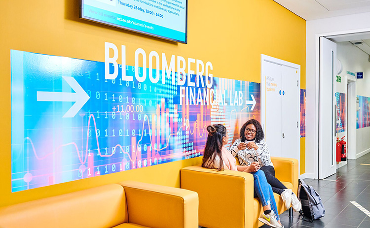 Two students sitting on a yellow sofa in front of a large colourful sign reading "Bloomberg Financial Lab" with an arrow pointing right