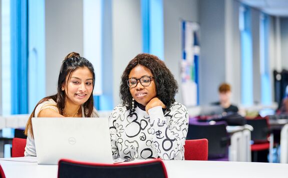 Two female students, one with light skin and long dark hair, and one with medium skin and curly black hair, sitting together at a table in a bright study space with a laptop.