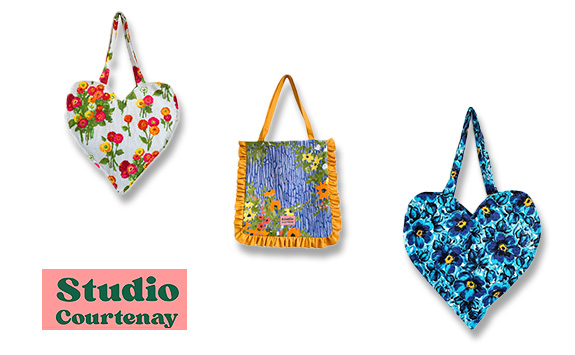 Three handmade fabric tote bags from Studio Courtenay displayed on a white background. Two are heart-shaped, one with a red floral print and the other with a blue floral pattern. The third bag is rectangular with a blue and yellow ruffled design.