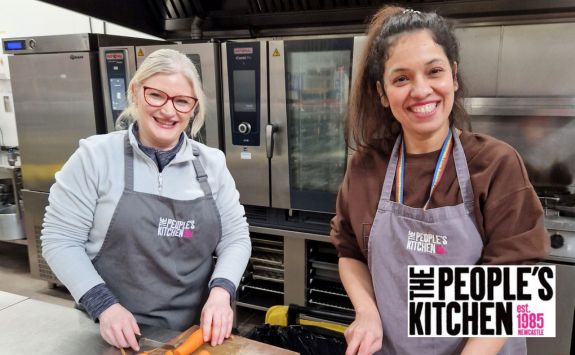 Two women smiling in a commercial kitchen setting, both wearing aprons with "The People’s Kitchen" logo. The woman on the left (Sue) has light skin, blonde hair, and glasses. The woman on the right (Sonia) has medium skin and dark hair.