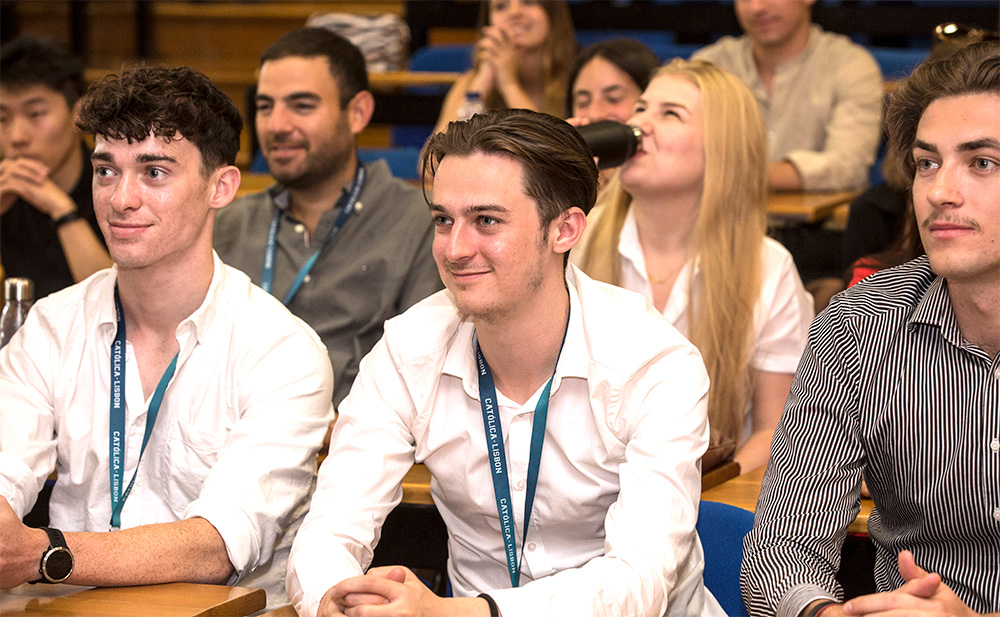 A group of students seated in a lecture hall at Católica Lisbon, engaged and listening attentively. The students are a mix of light-skinned and medium-skinned individuals, wearing business-casual attire, with university lanyards around their necks.