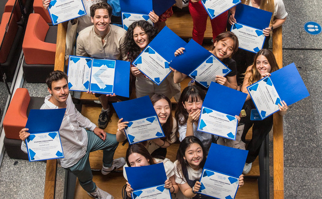 A diverse group of summer school students celebrating, holding blue and white certificates while standing in an atrium. The group includes male and female students, with some smiling and others looking up at the camera.