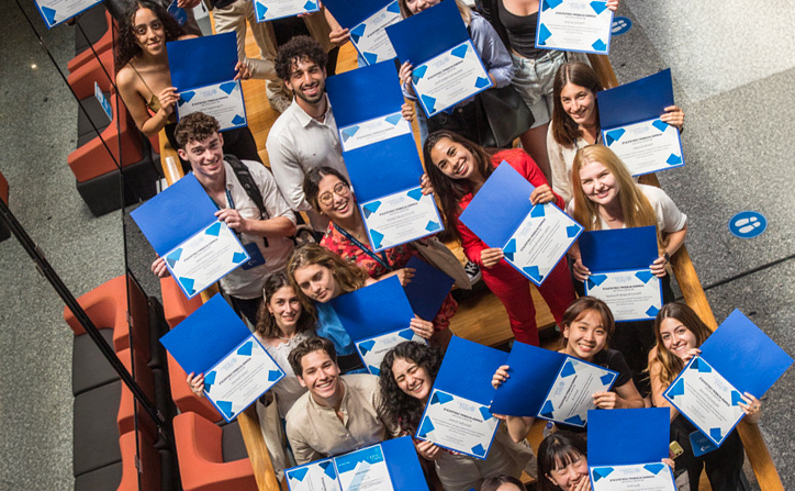 A group of diverse summer school students celebrating, holding blue and white certificates while standing in an atrium. The group includes light-skinned, medium-skinned, and dark-skinned individuals, with some smiling and others looking up at the camera.