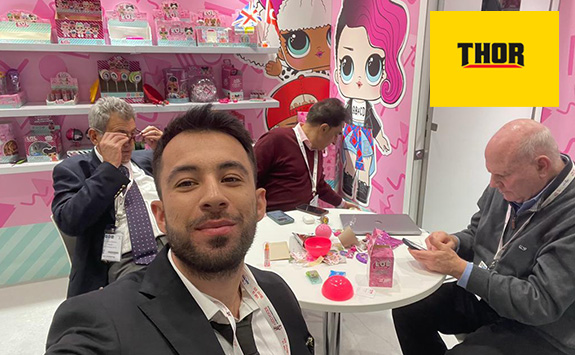 Ilker Aybey, founder of Thor Teknoloji, takes a selfie in a vibrant exhibition booth featuring colourful toy displays and branding. He is wearing a black suit and white shirt. Three other people, two older men and one middle-aged man, sit at a round table behind him. The yellow Thor Teknoloji logo is visible in the top-right corner.