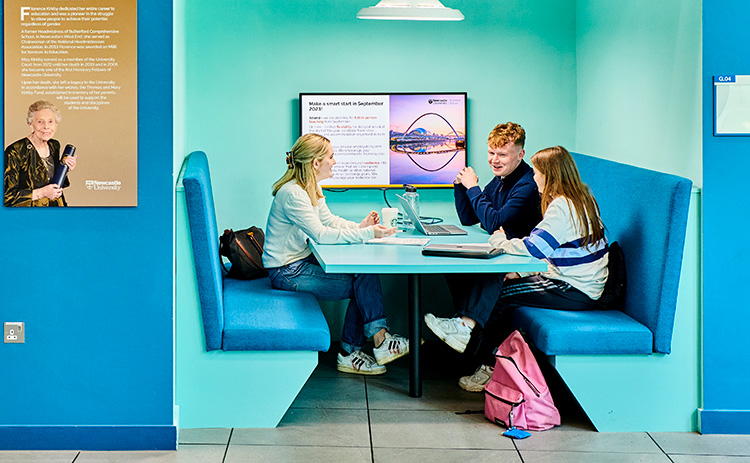 Three students sitting in a booth, working together on laptops, with a presentation screen on the wall behind them in a brightly coloured study space