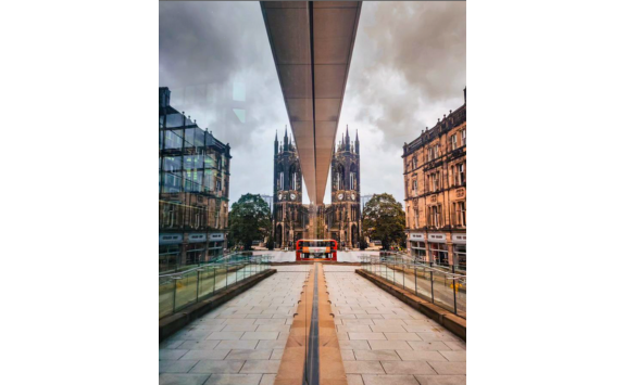 Winning entry of Welcome Hyem competition 2023 by "@i_am_tarkik" - a reflective view of Newcastle upon Tyne featuring a historic cathedral, symmetrical modern architecture, and a red bus on a cloudy day.