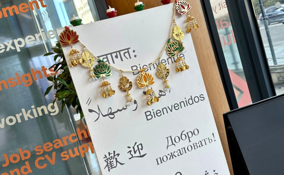 A decorative welcome sign adorned with golden ornaments and garlands, displaying greetings in various languages including English, Spanish, Arabic, Chinese, Russian, and more. The sign stands next to a glass wall with bold orange text