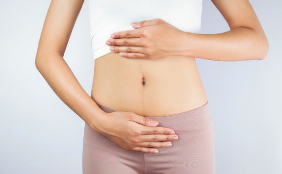 Light-skinned woman wearing a white crop top and pink trousers holding her abdomen with both hands,