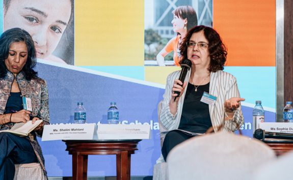Cristina Neesham speaking at a formal event. She is holding a microphone and engaging with the audience. Next to her, another woman is seated and taking notes.