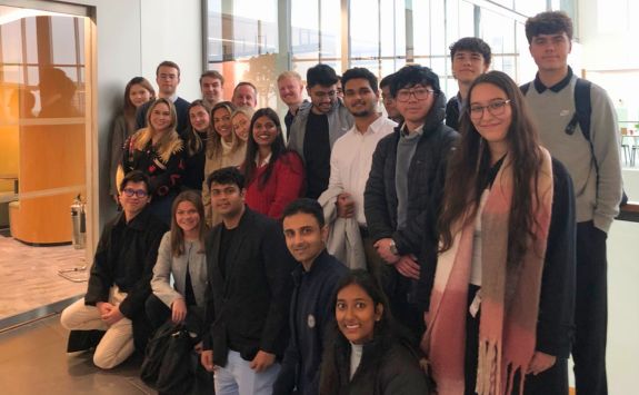 A group of students posing for a photo during the student trek. They are indoors, standing near a glass-walled area with a modern office background.