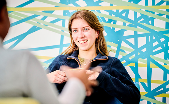 A student sitting in the Urban Cafe, in the Urban Sciences Building, drinking a coffee and talking with a colleague. 