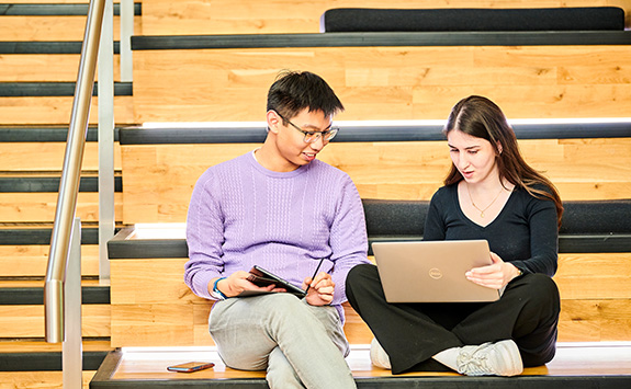 Two students sitting together on the stairs, one with a laptop.