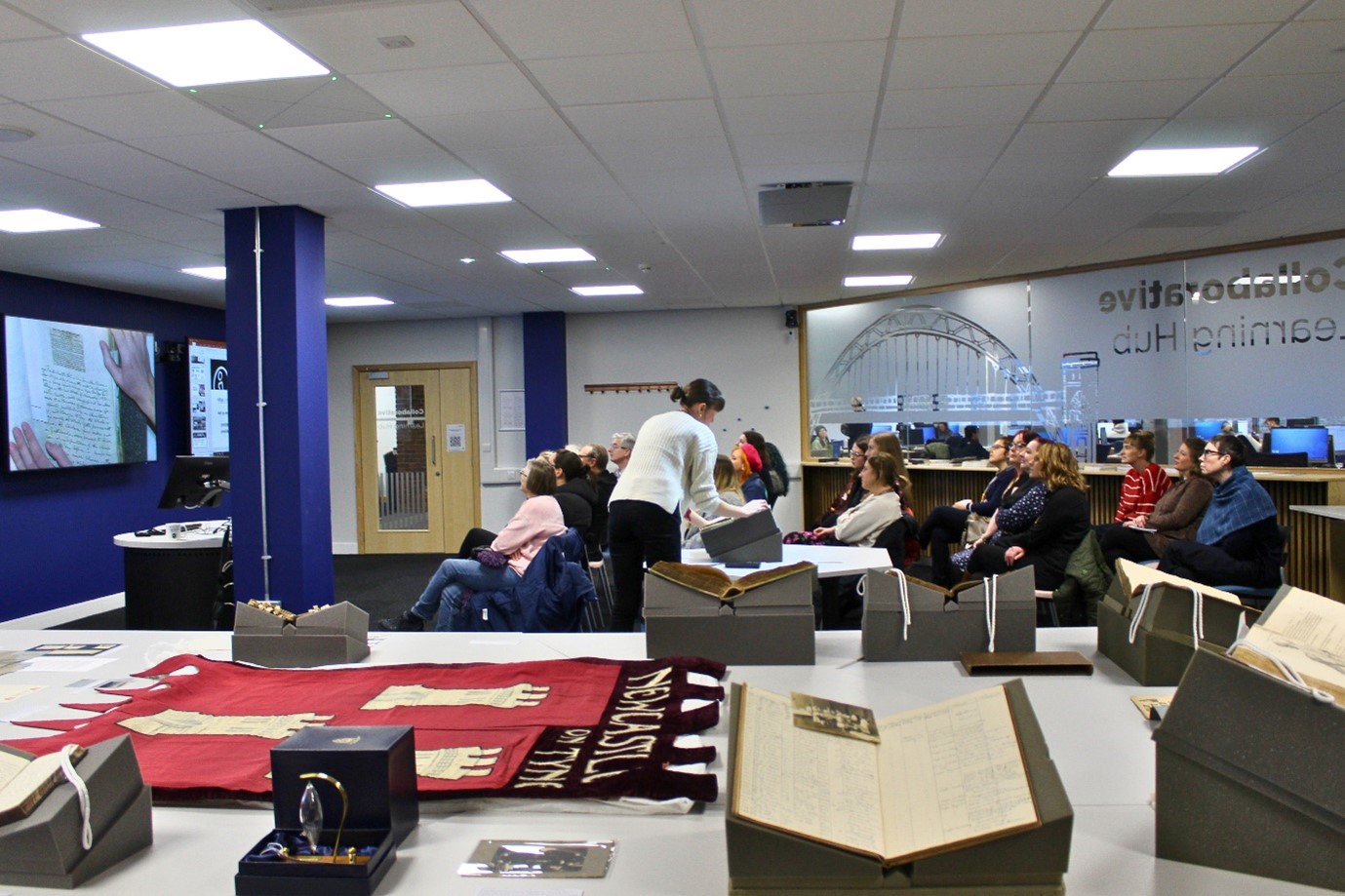 Photo of people sat in a room looking at an item being projected onto screens, with items on display from Special Collections & Archives, within the Collaborative Learning Hub space