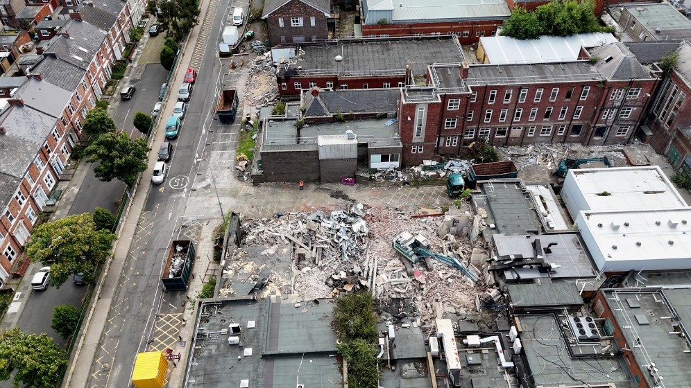 An aerial view of the western edge of the HIN site. Two excavators can be seen demolishing disused hospital buildings. There are large skips and waste lorries waiting to receive the demolished materials. Demolition has only started recently and so this image shows some demolition but lots of other buildings still standing.