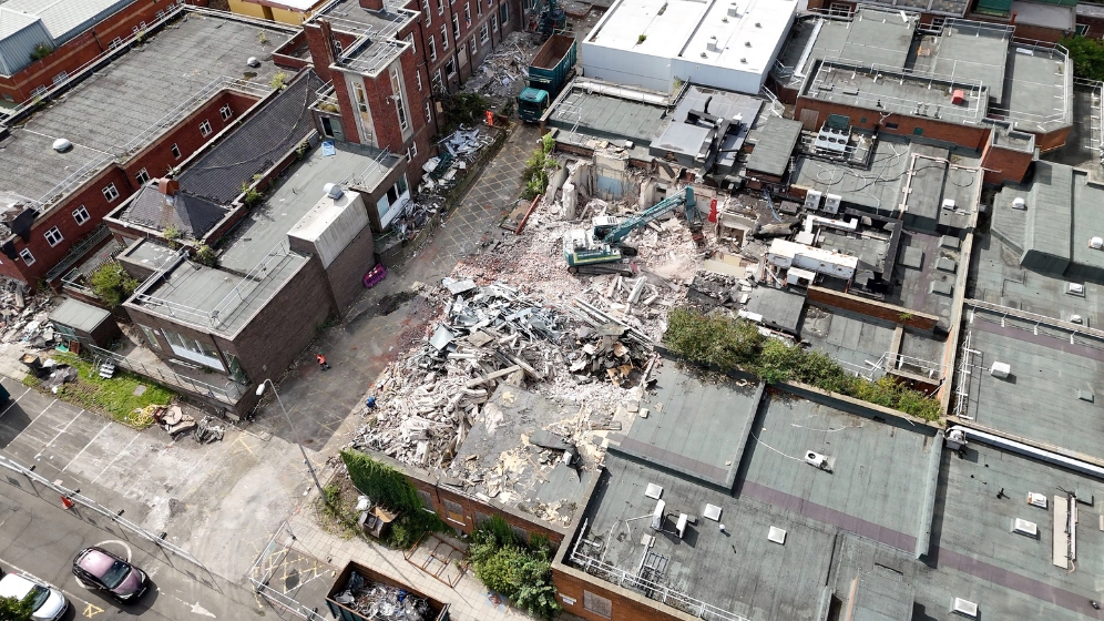 An aerial view of the western edge of the HIN site. An excavator can be seen demolishing disused hospital buildings. There are large skips and waste lorries waiting to receive the demolished materials. Demolition has only started recently and so this image shows some demolition but lots of other buildings still standing.